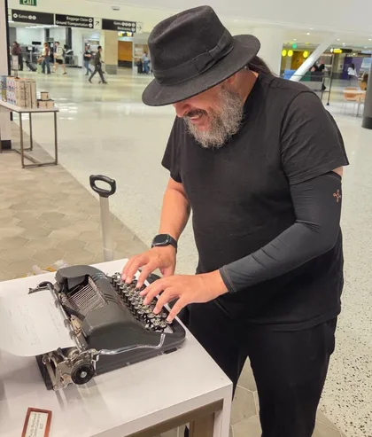 Hassan, in a black hat, grinning while typing on an old manual typewriter in an airport terminal